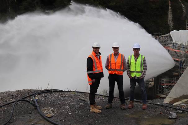 A group of men wearing safety vests and helmets standing in front of a large water spout

Description automatically generated